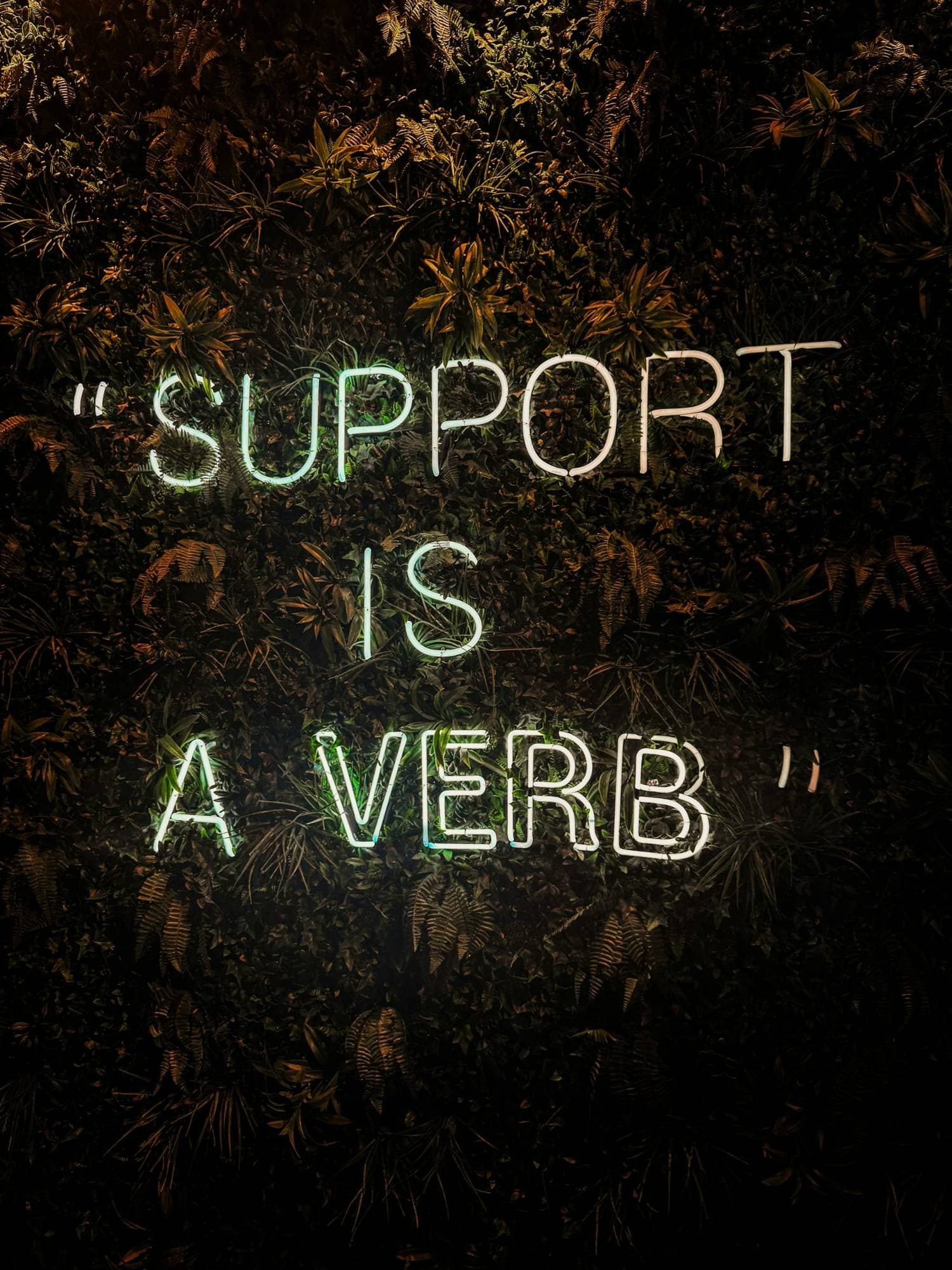 Neon sign reading Support Is A Verb on a dark plant wall